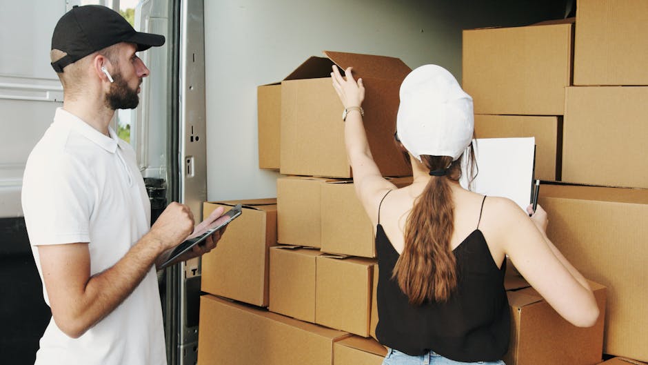A man and a woman are standing beside a van during a home relocation process, surrounded by stacks of cardboard boxes of various sizes, some open and partially filled with packing materials. The woman, wearing a white cap and black sleeveless top, is reaching up to a large box, possibly for arranging or inspecting its contents, while holding a clipboard in her left hand. The man, dressed in a white polo shirt and black cap, is holding a tablet or clipboard and appears to be overseeing or documenting the packing and moving process. The scene takes place at the back of a moving van, with a pavement visible in the background. Both individuals are engaged in loading or organizing boxes, aided by equipment such as straps or blankets for furniture protection, suggesting an organized furniture transport and packing service by Man and Van Tottenham, supporting efficient home removals and logistics.