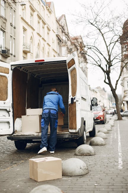 A man wearing a blue hoodie and dark trousers is inside the rear cargo area of a white delivery van parked on a city street, preparing packed items for a home relocation. The van's interior walls are lined with wooden panels, and several cardboard boxes and foam packing materials are visible inside. One cardboard box rests on the cobblestone pavement directly behind the van, while the vehicle is positioned close to a row of large, rounded street bollards lining the sidewalk. In the background, there are multi-storey buildings with white facades and window balconies, along with a leafless tree and parked vehicles, including a red car and a black van. The scene takes place during daytime under overcast skies, capturing a typical moment in furniture transport and packing for professional removals by Man and Van Tottenham, emphasizing the loading process of household items in preparation for house removals.