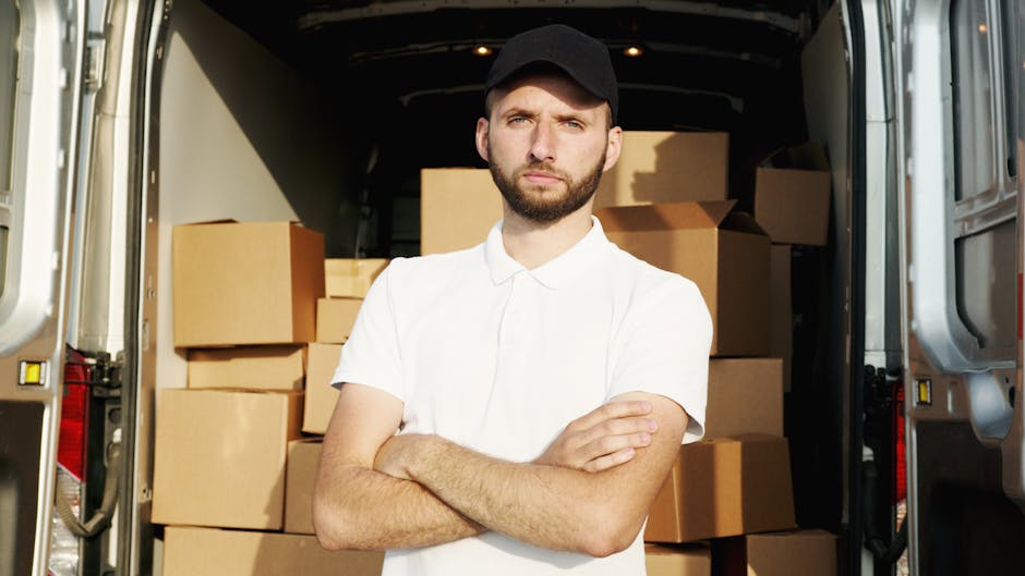 A young man with light brown hair and a short beard is sitting on the edge of a van's open cargo area during a home relocation. He is dressed in a blue zip-up hoodie, dark blue trousers, and white sne