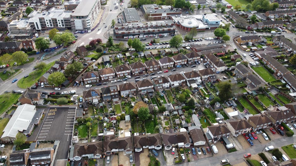 An aerial view of a residential neighbourhood in Tottenham, showing rows of semi-detached houses with small garden spaces, some featuring solar panels on their roofs. The houses are aligned along narrow streets with parked cars on driveways and curbs. Behind the row of houses, there are commercial buildings and a large parking lot with numerous vehicles. The scene includes green trees and well-maintained lawns, with clear weather illuminating the area. This image captures the typical layout of houses involved in home relocation or furniture transport processes, relevant to house removals and moving logistics offered by Man and Van Tottenham, as seen on their webpage for Tottenham Hale removals in Hale Village flats.