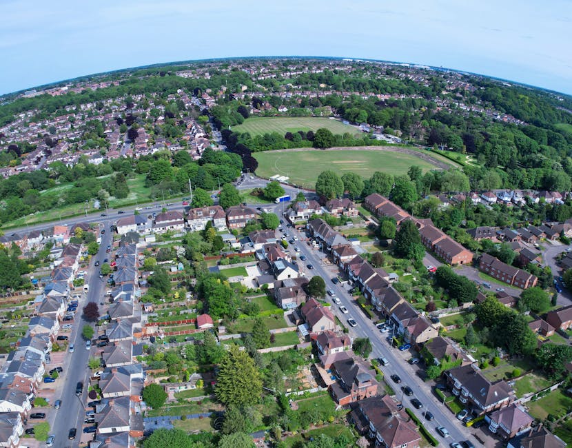Aerial view of a residential neighbourhood in Tottenham Hale, showing rows of terraced houses with pitched roofs, front gardens, and parked cars along the streets. Behind the houses, there are large green areas with open sports fields and scattered trees, indicating a community park or recreational space. The landscape extends towards the horizon, with a mixture of trees, residential buildings, and suburban development under a clear sky. Visible within the scene is a busy street in the foreground, with vehicles lining the road, consistent with a typical home relocation environment. The image highlights the urban and green spaces of Tottenham Hale, often involved in furniture transport, packing, and loading during removals, as coordinated by companies like Man and Van Tottenham for their Tottenham Hale removals guide for Hale Village flats.