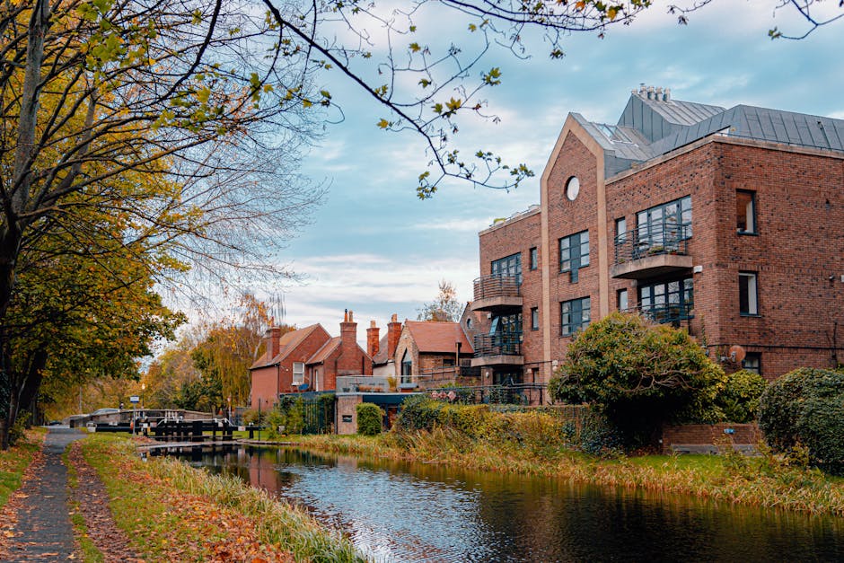 An aerial view of a residential neighbourhood in Tottenham, showing rows of semi-detached houses with small garden spaces, some featuring solar panels on their roofs. The houses are aligned along narrow streets with parked cars on driveways and curbs. Behind the row of houses, there are commercial buildings and a large parking lot with numerous vehicles. The scene includes green trees and well-maintained lawns, with clear weather illuminating the area. This image captures the typical layout of houses involved in home relocation or furniture transport processes, relevant to house removals and moving logistics offered by Man and Van Tottenham, as seen on their webpage for Tottenham Hale removals in Hale Village flats.